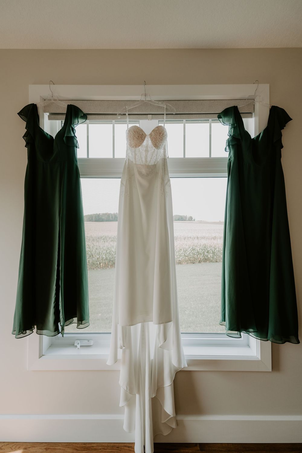 hanging in window two forest green bridesmaid dresses with the bride's dress in the centre