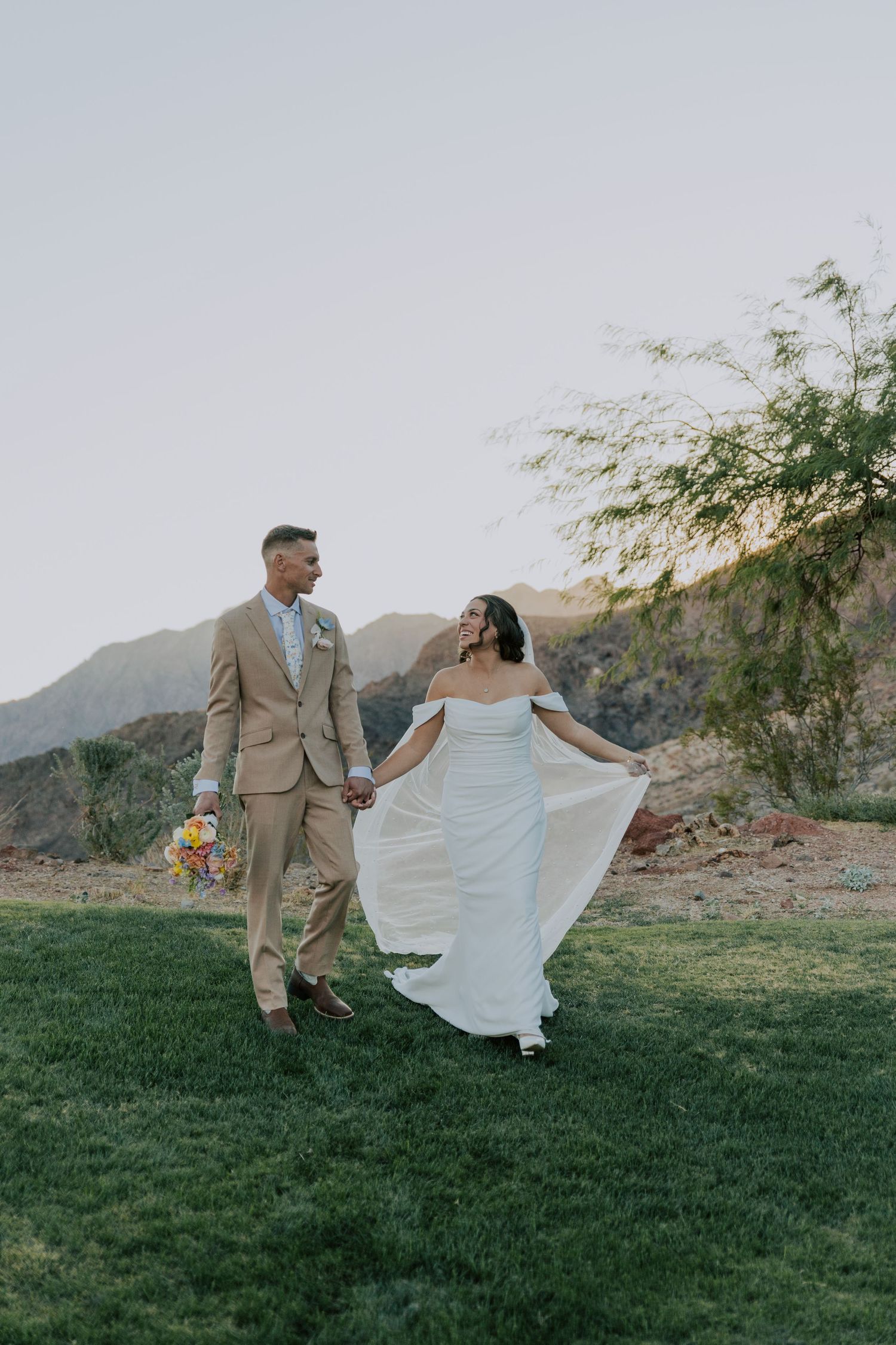 Bride and groom holding hands walking on green lawn with mountain backdrop at sunset.