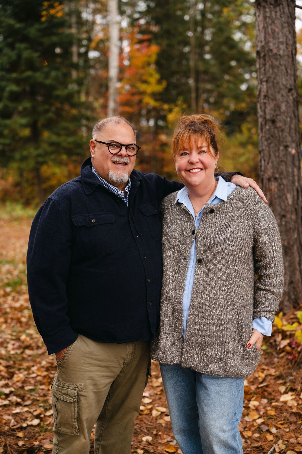 Couple embraces while standing on autumn leaves in a wooded forest setting.