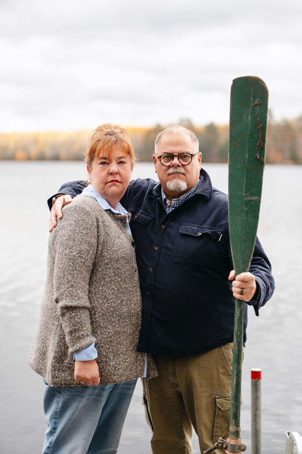A couple poses playfully together by a wooden post near a lake on an autumn day.
