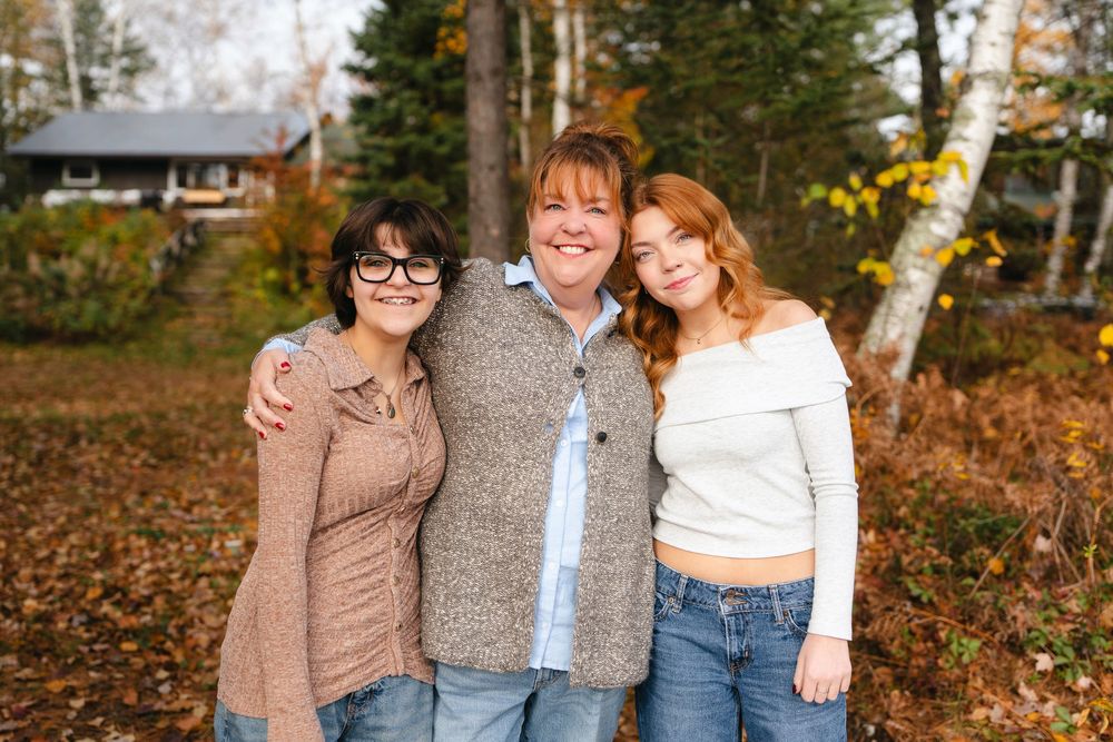 Three people stand together outdoors in casual fall clothing with autumn foliage and a cabin in the background.
