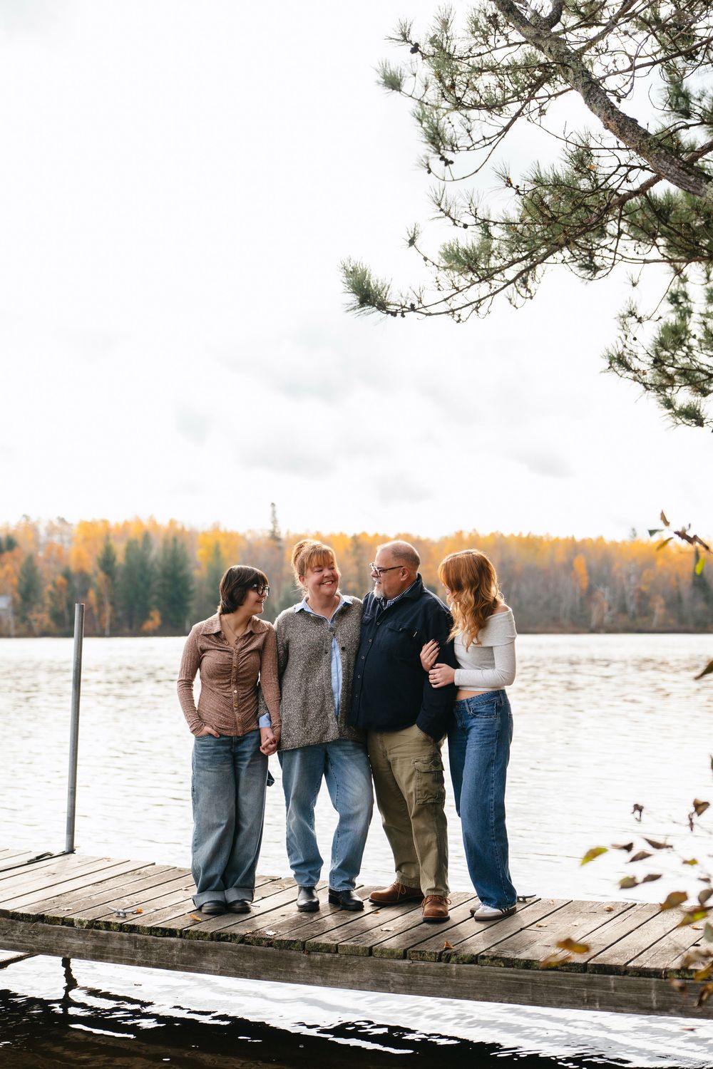 People stand together on a wooden pier overlooking a serene lake with fall foliage in the background.