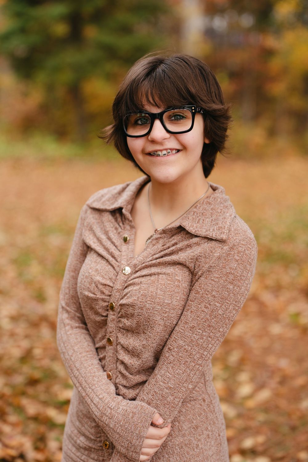 Person wearing glasses and a brown sweater smiles playfully in autumn setting with fallen leaves.