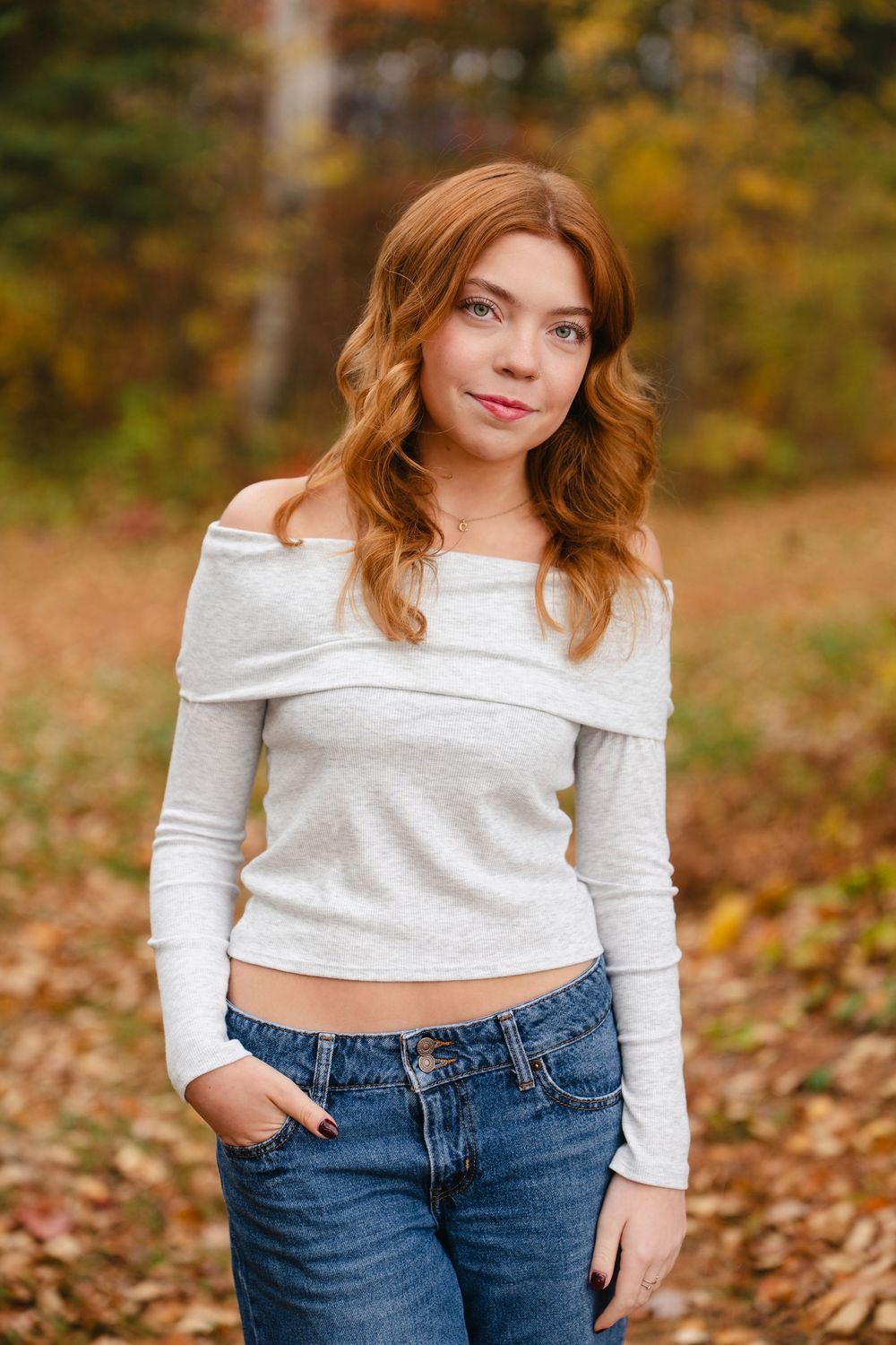 Person in white off-shoulder top and jeans poses against autumn background with fallen leaves.
