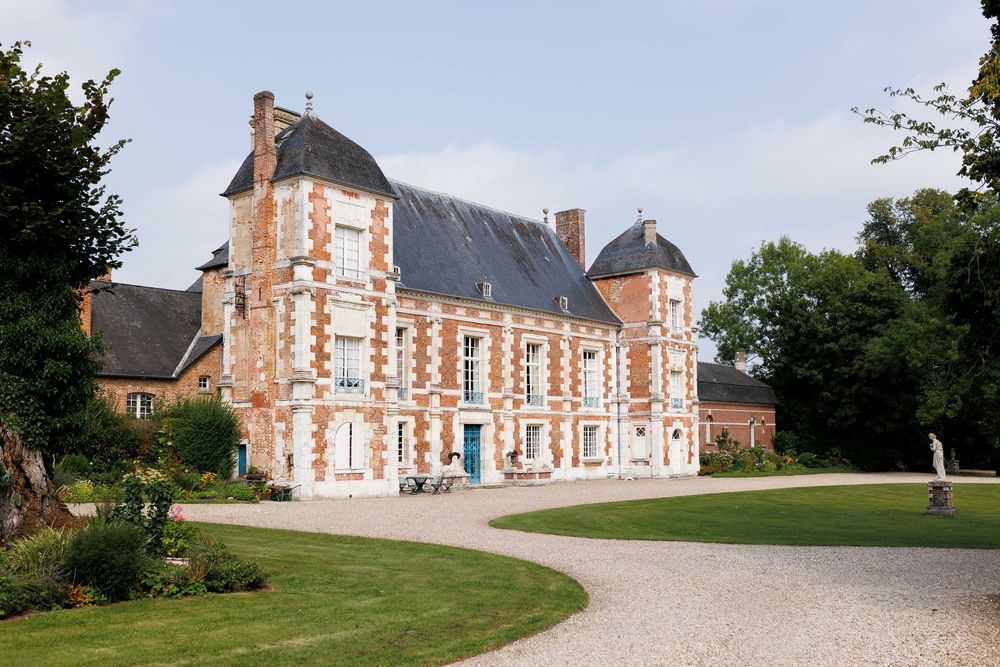 Panoramic view of a grand French chateau with red brick and stone architecture surrounded by manicured lawns and gardens.