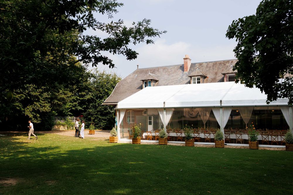 A white wedding tent set up on a lush green lawn next to a thatched-roof cottage on a sunny summer day.