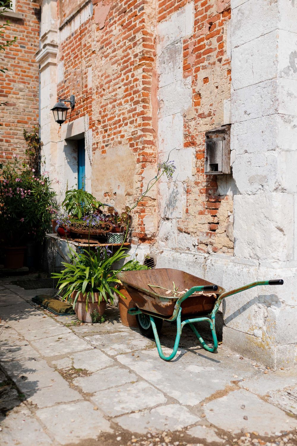 Rustic turquoise wheelbarrow against weathered brick wall with exposed plaster and stone pavement in vintage setting.