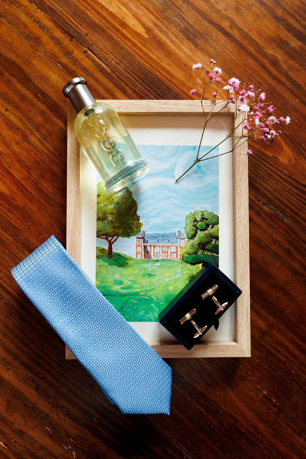 Wedding accessories including blue tie, cologne bottle and painting of mansion venue arranged on wooden surface with pink flowers.