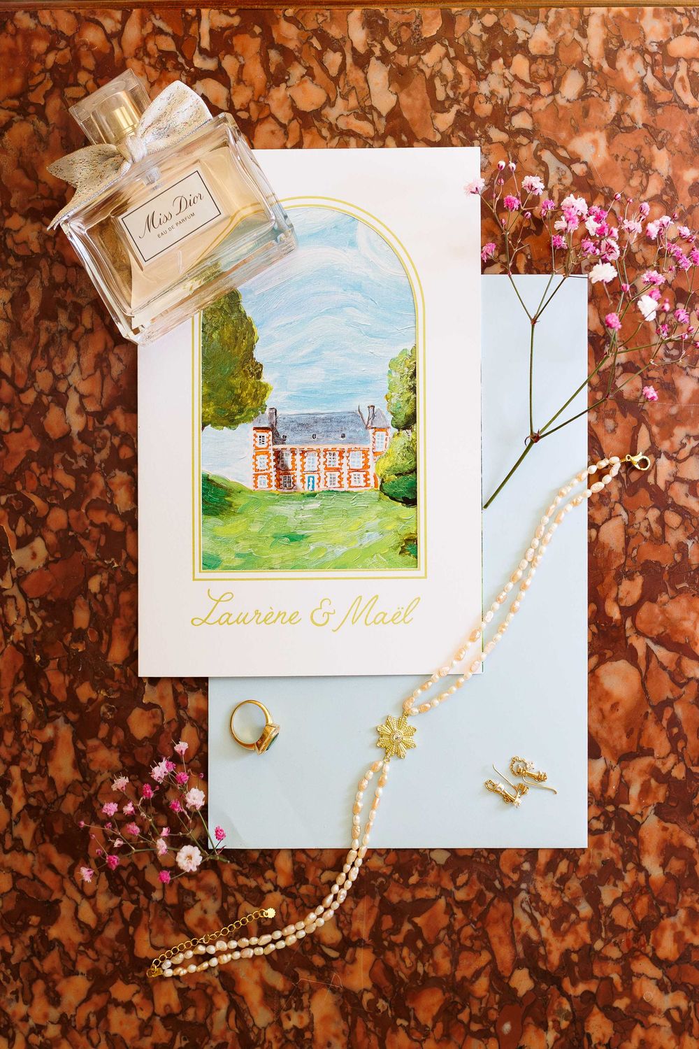 Wedding details featuring watercolor venue illustrations, pearl necklaces, and perfume bottles arranged on a rustic brown background.