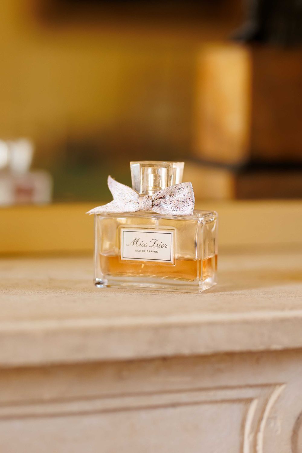 A glass perfume bottle with pink liquid sits on a beige surface in soft lighting.