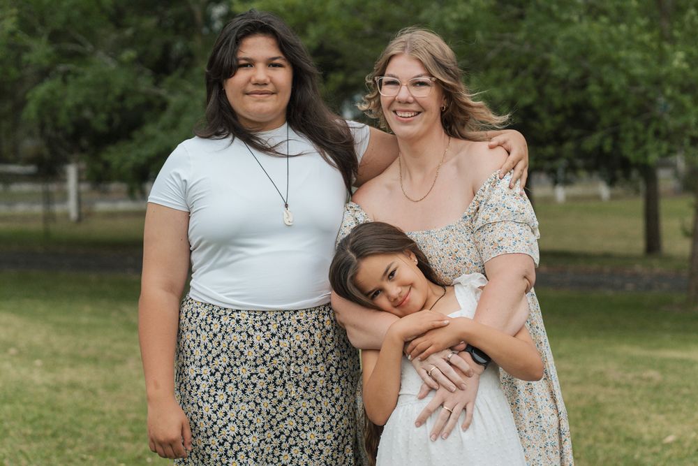 Three people embrace and smile together outdoors in a park setting on a sunny day Box Hill The Gables
