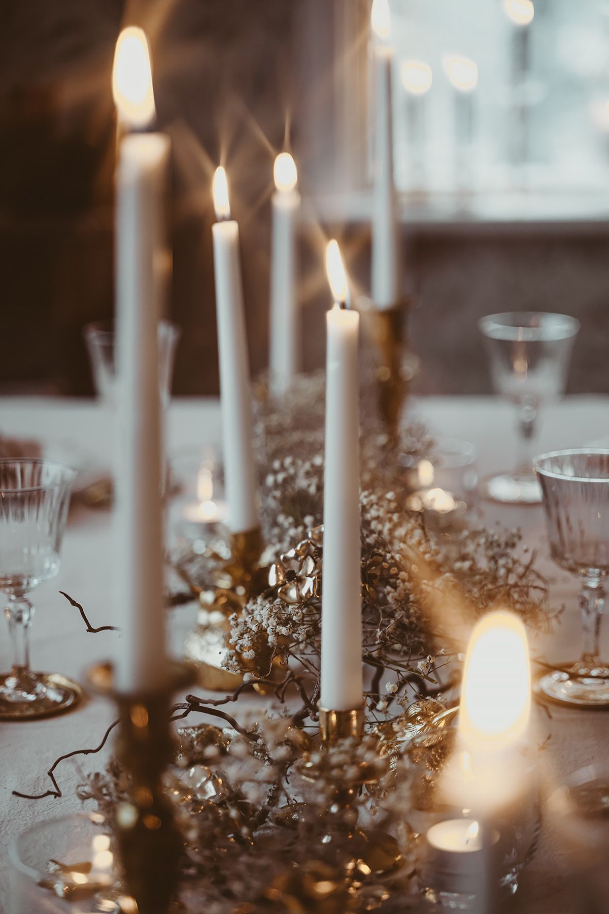 Dekorativt uppdukat bord i vitt och guld med vita levande ljus och kristallglas. På bordet ligger torkade växter och brudslöja.