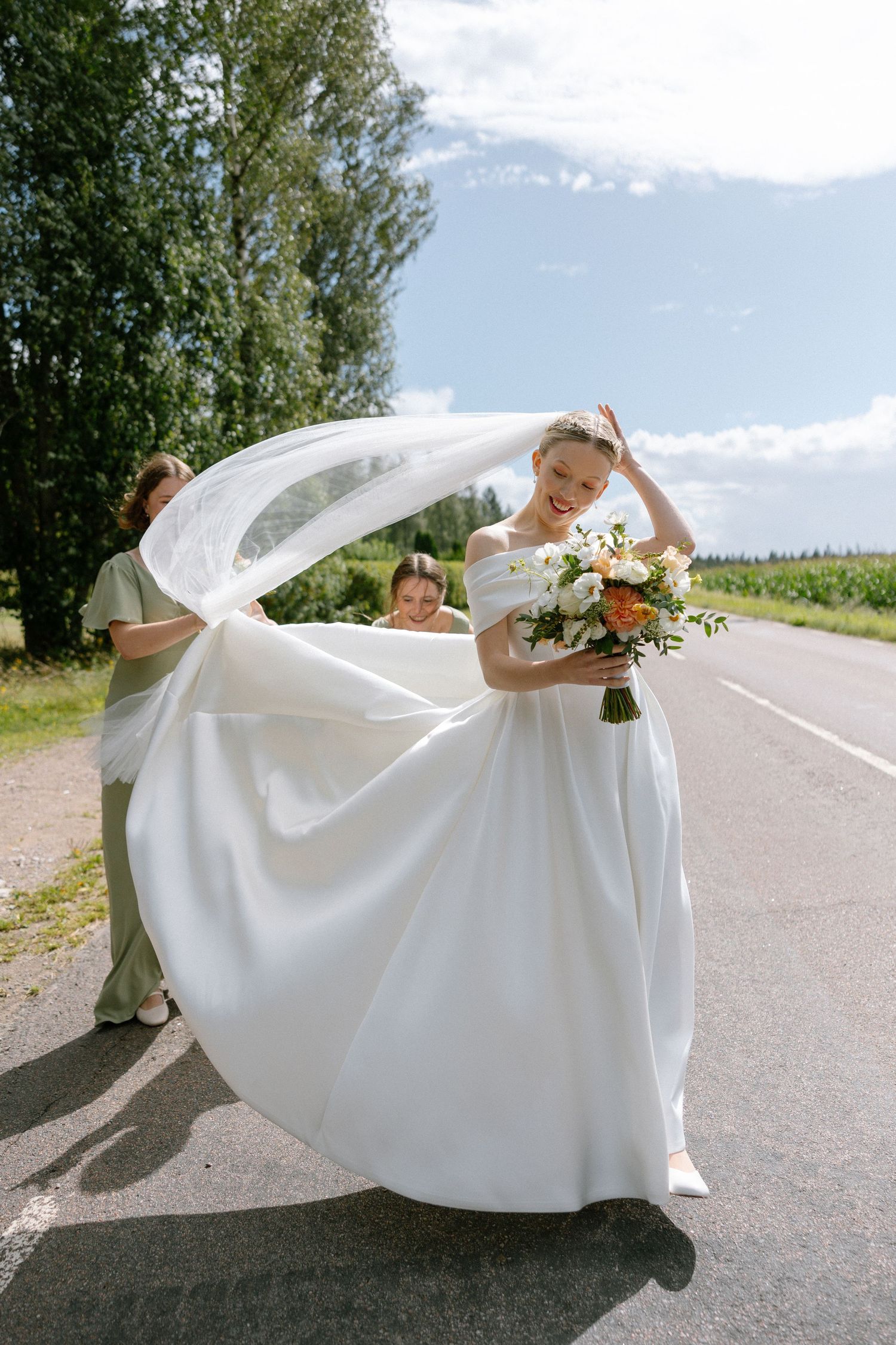 A flowing white wedding dress billows in the wind on a scenic mountain road during a summer photoshoot.