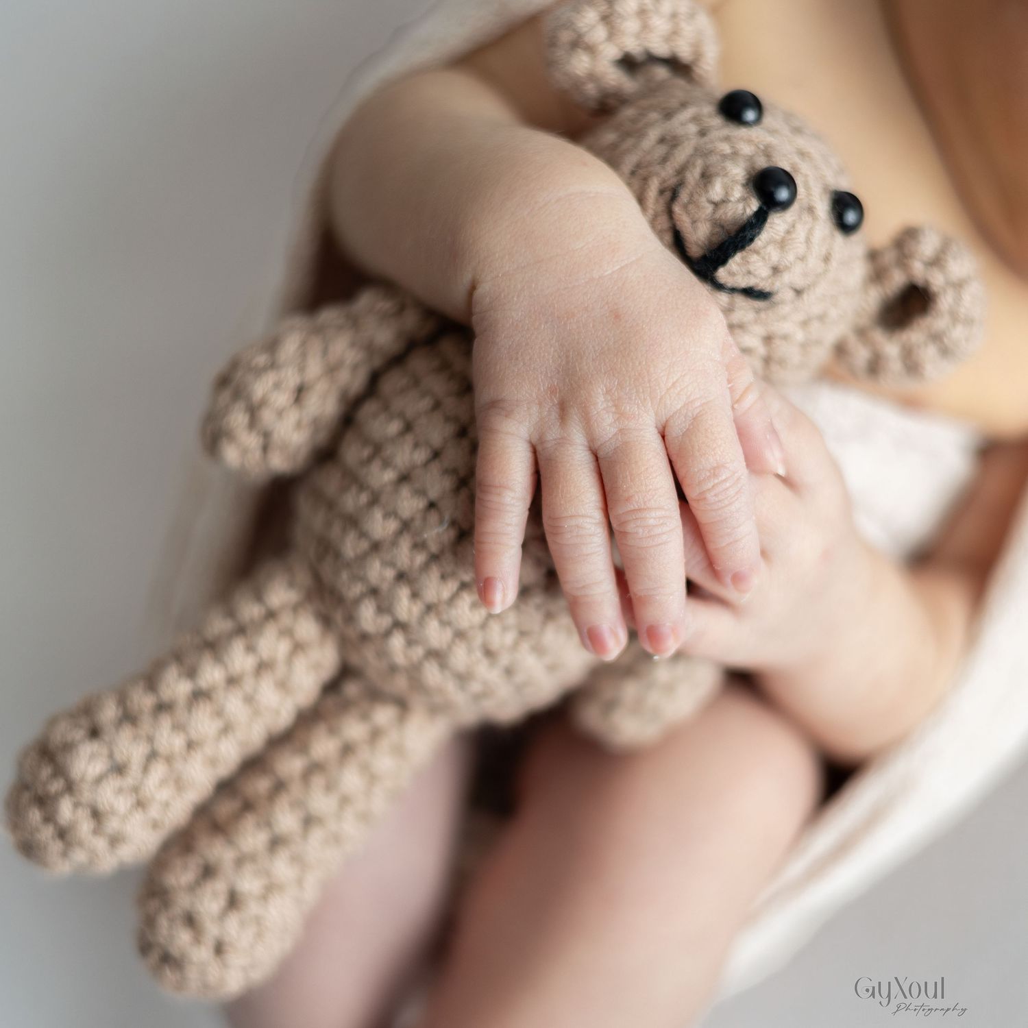 A tiny hand gently holds onto a knitted beige teddy bear in a soft, intimate close-up photo.