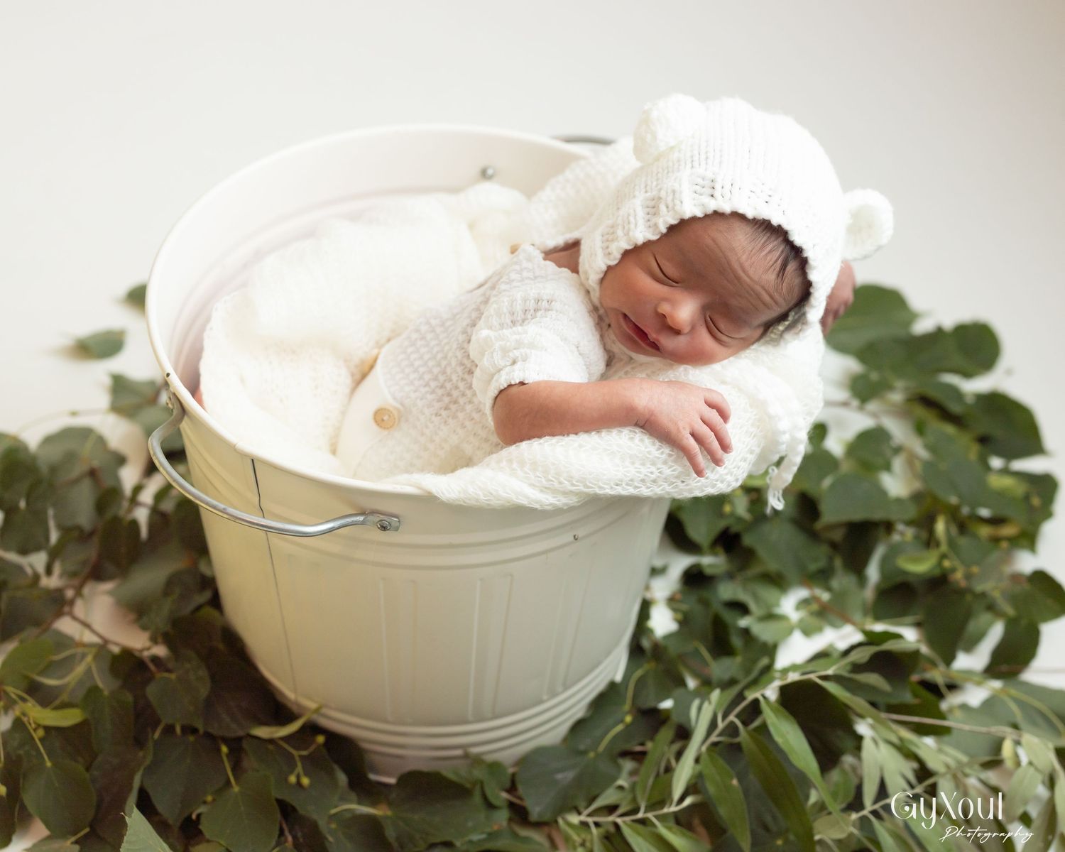 A sleeping newborn baby wrapped in white blankets rests in a cream-colored bowl surrounded by green eucalyptus leaves.