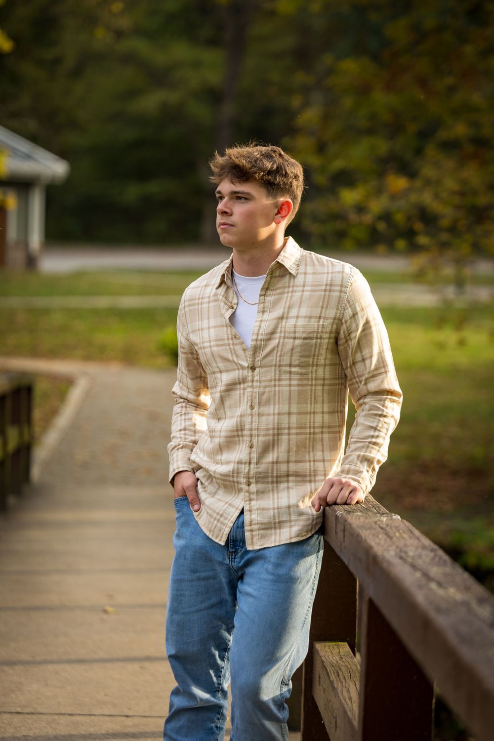 Person in plaid button-down shirt and jeans leans against wooden railing in outdoor autumn setting.