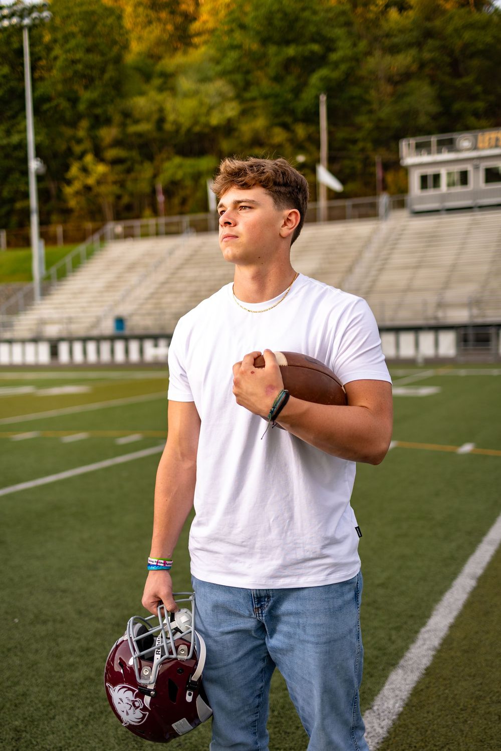 Football player stands on field holding helmet while wearing white t-shirt and jeans during golden hour.