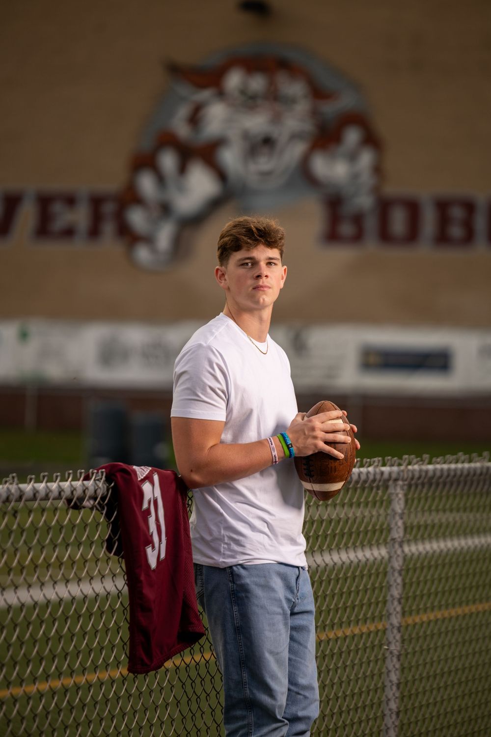 Person in white t-shirt and jeans stands by chain link fence holding football during sunset.