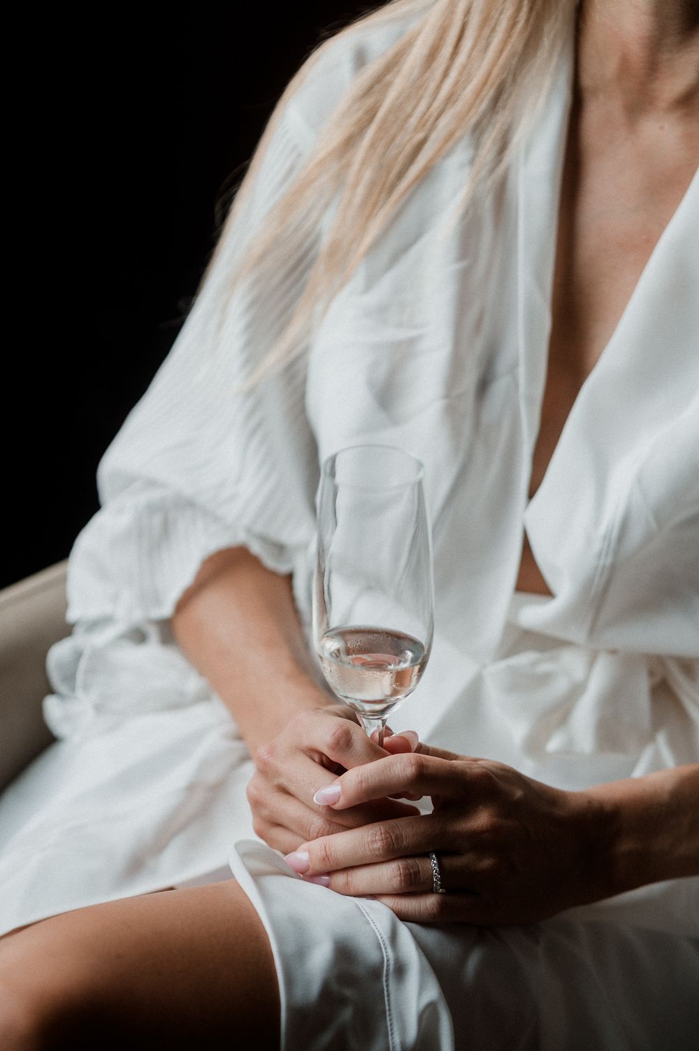 Close-up shot of hands holding a delicate wine glass in a white silk robe against a dark background.
