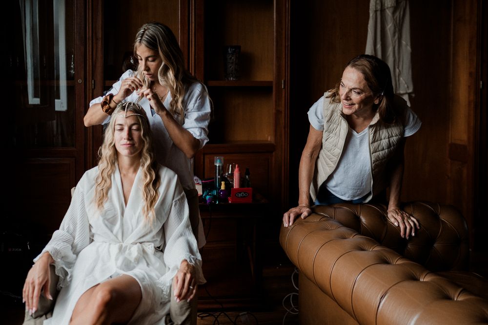 A group of friends in white clothing laugh together while sitting on a brown leather couch in a dimly lit room.