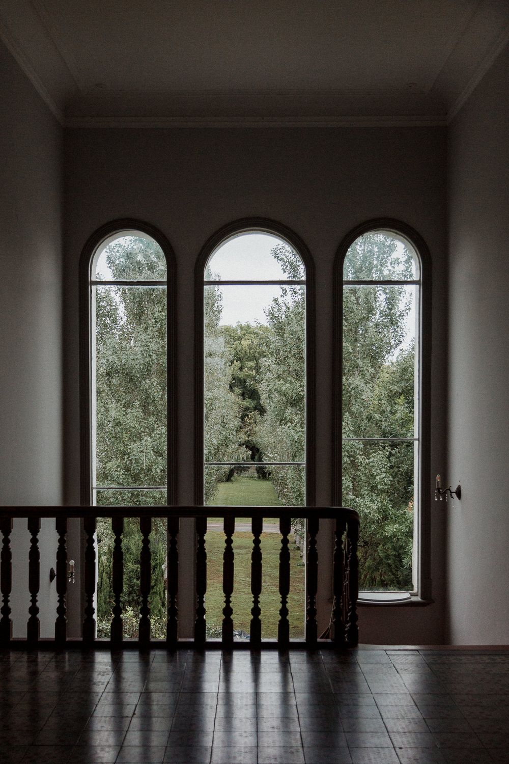 Three arched windows with dark wooden railings frame a view of trees in a moody interior hallway setting.
