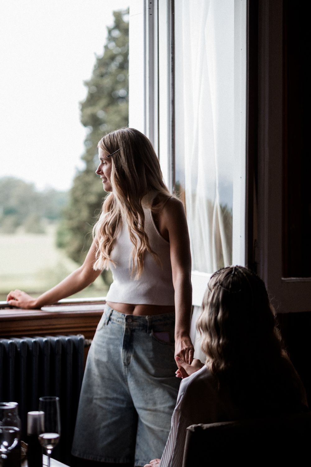 A woman in a white top and jeans stands by a window in a restaurant, creating a serene silhouette effect.