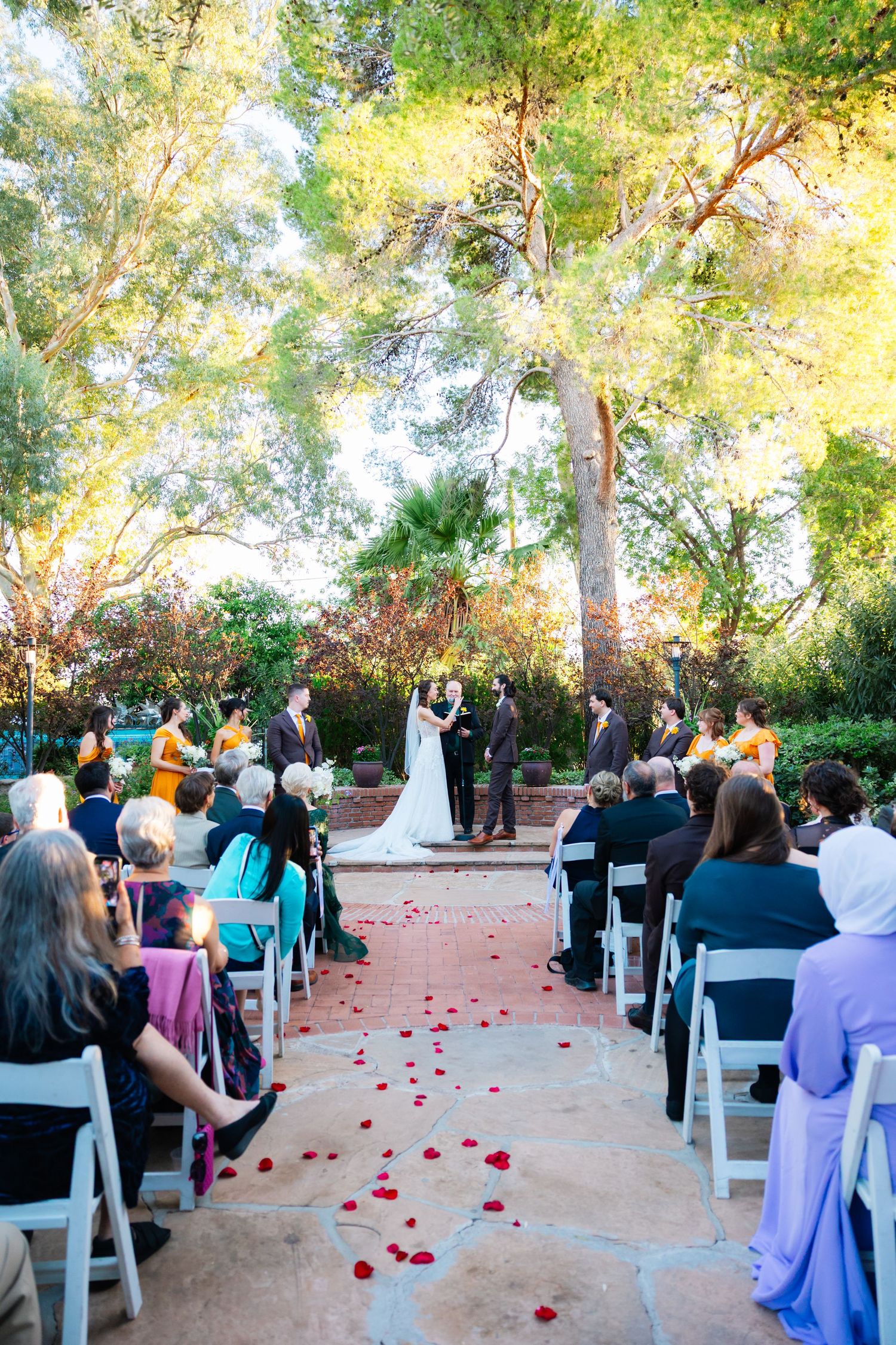 Outdoor wedding ceremony setup with white chairs and rose petals scattered along a brick paved aisle.