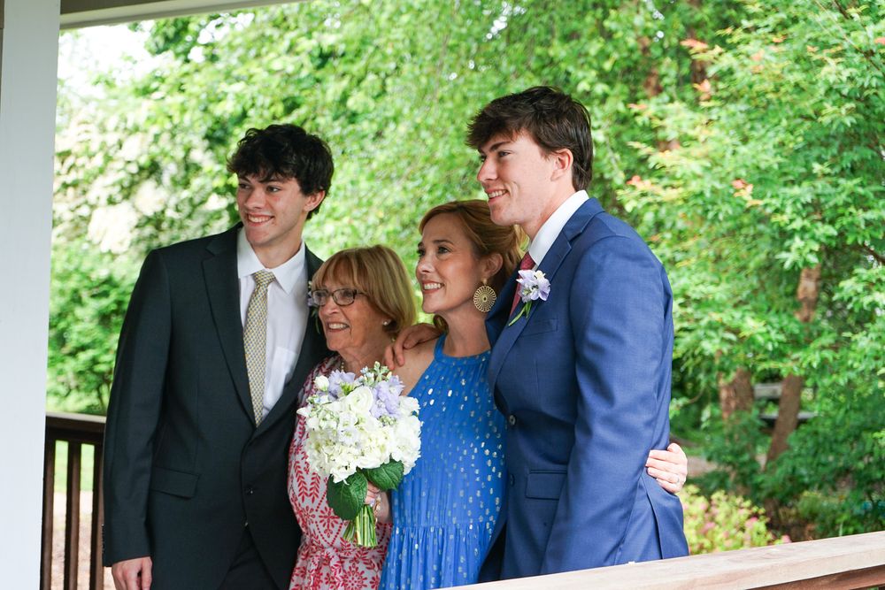 Wedding party members pose together on a covered outdoor deck surrounded by lush green trees in summer.