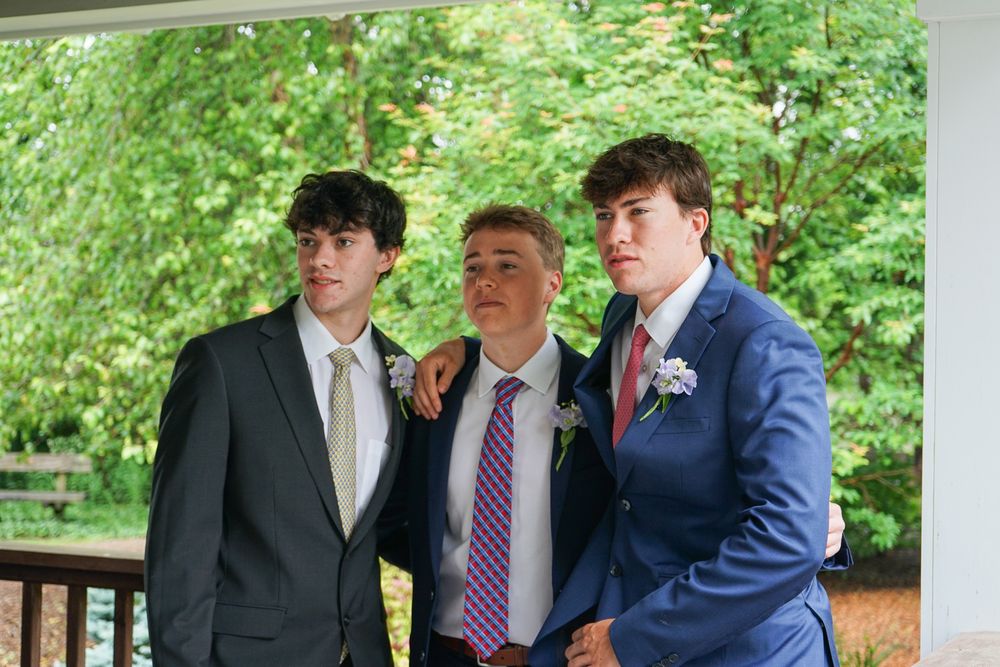 A group in formal attire stands together on a covered porch surrounded by lush green foliage in the background.