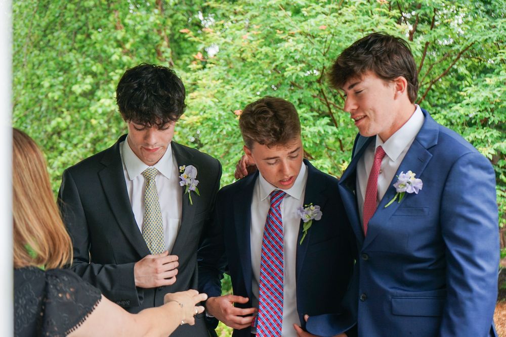 Wedding party members in formal attire gather on a white porch surrounded by green trees.