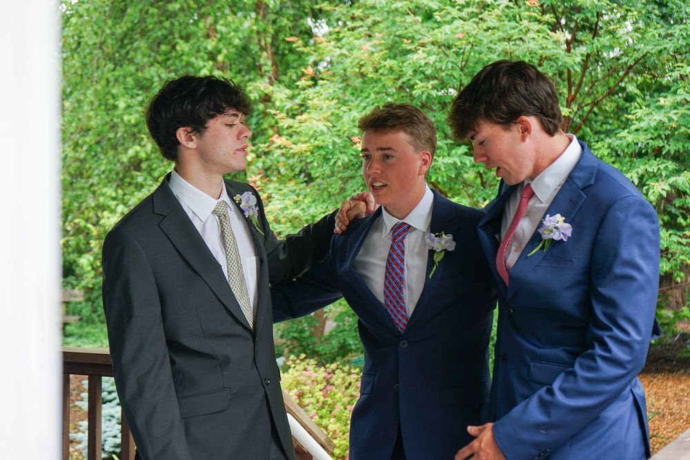 People in formal attire gather outdoors on a porch with lush green foliage in the background.