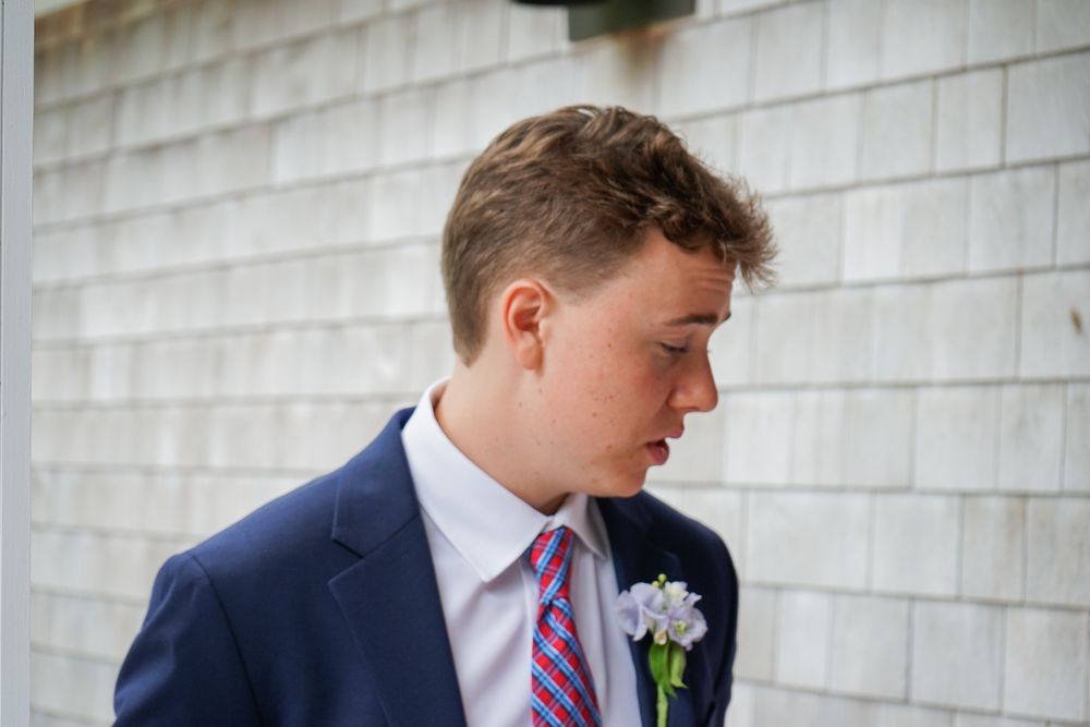 Young man in navy suit and pink striped tie poses formally against white siding.
