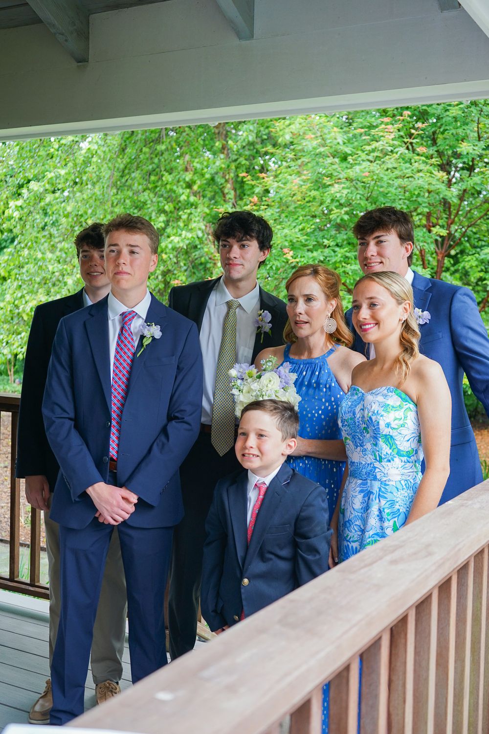 Wedding party members line up on a wooden deck wearing formal attire.