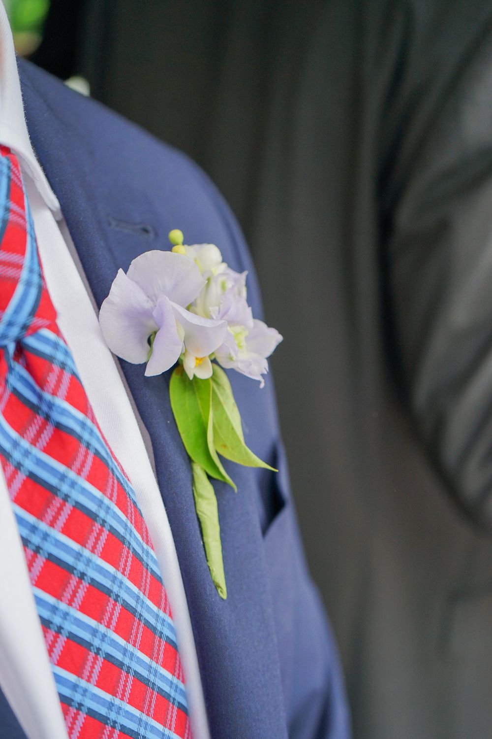 A white floral boutonniere pinned to a navy suit jacket with a red and blue plaid tie.