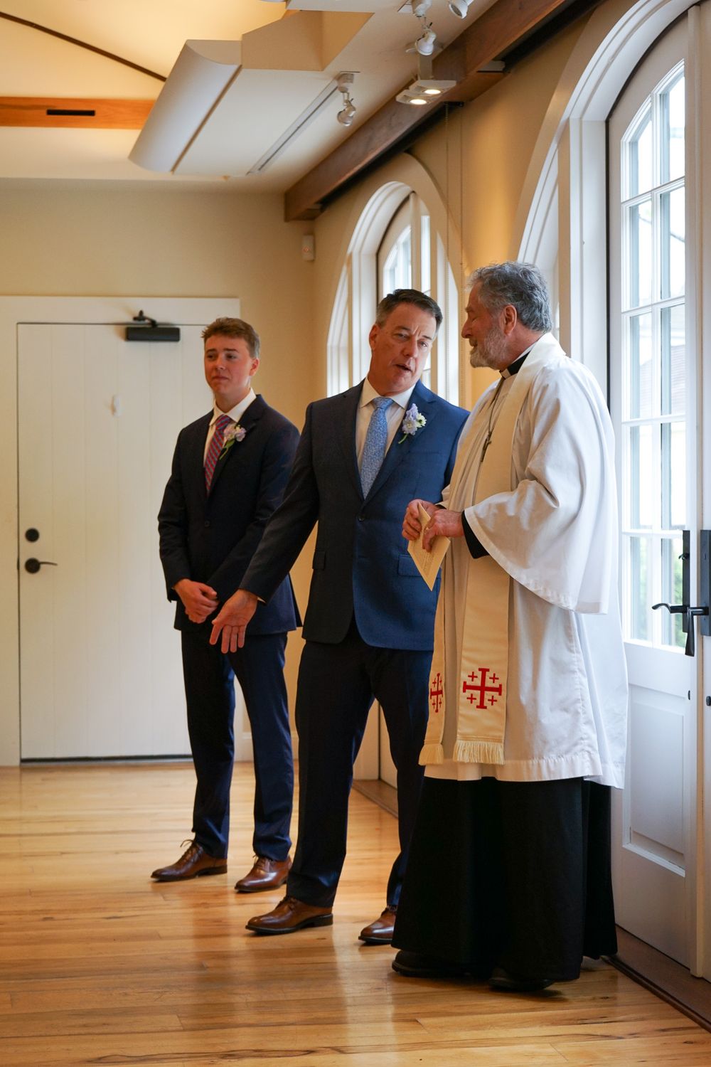Several people in formal attire stand in a bright room with wooden floors and white walls near a religious symbol.