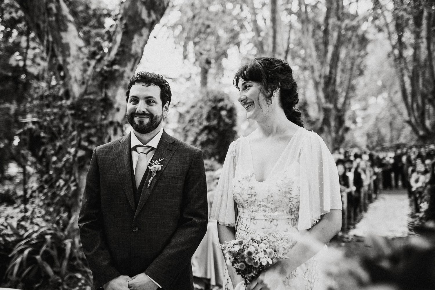 Black and white wedding portrait showing couple walking through wooded outdoor setting.