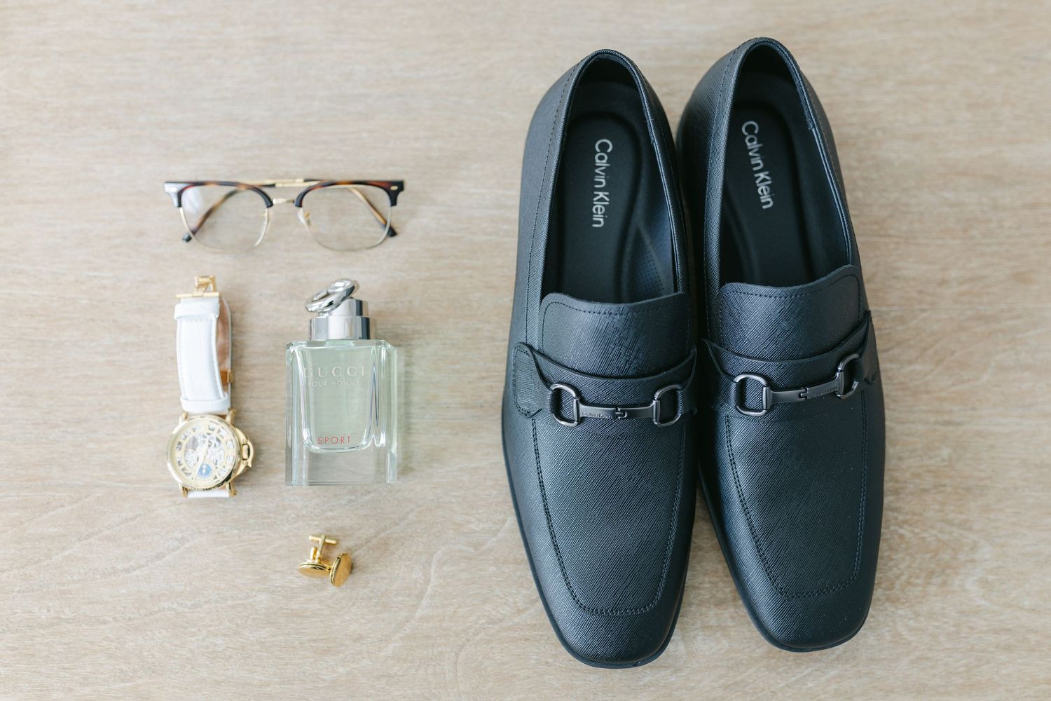 A collection of fashion accessories including black loafers, eyeglasses, perfume bottle, and jewelry on a beige surface.