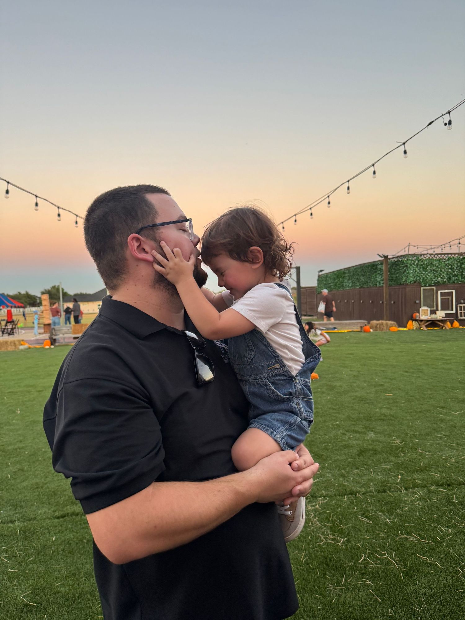 A tender moment captured at sunset on a grassy lawn with string lights overhead during an outdoor gathering.