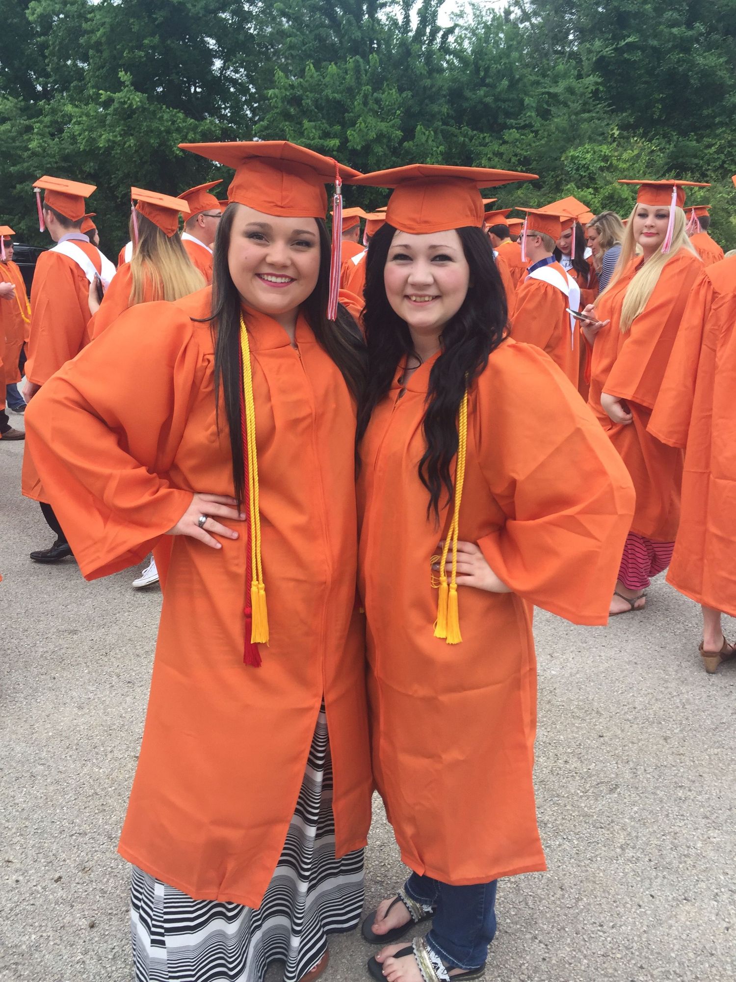 Two graduates in bright orange caps and gowns pose together at an outdoor commencement ceremony.