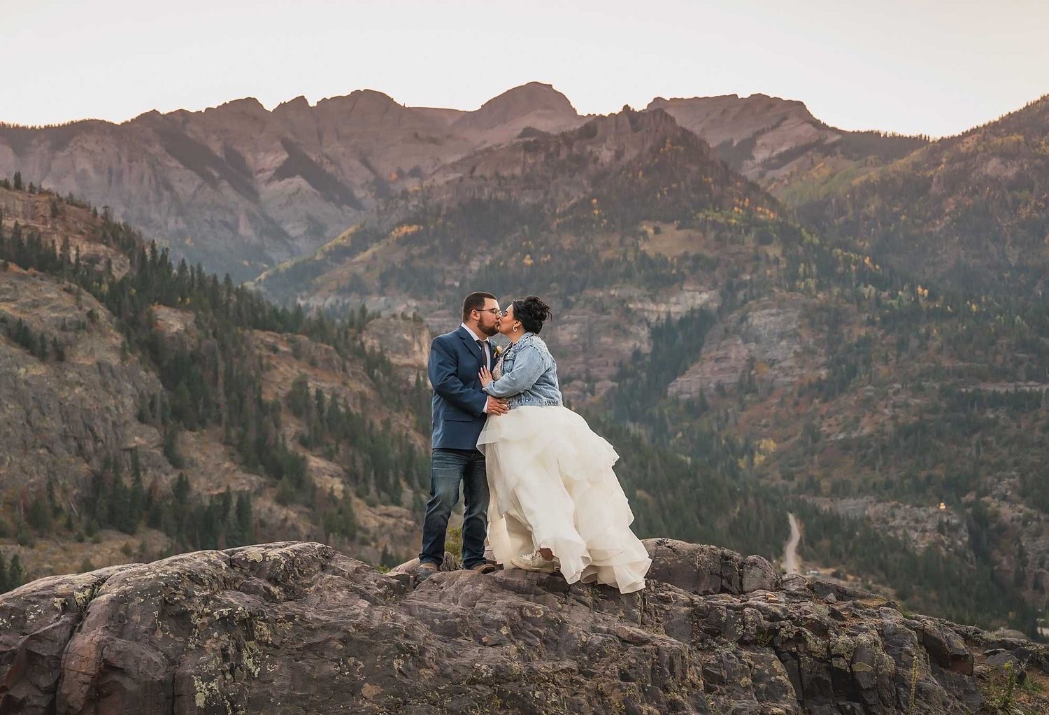 Couple in wedding attire embraces on mountain summit overlooking rugged peaks at sunset.