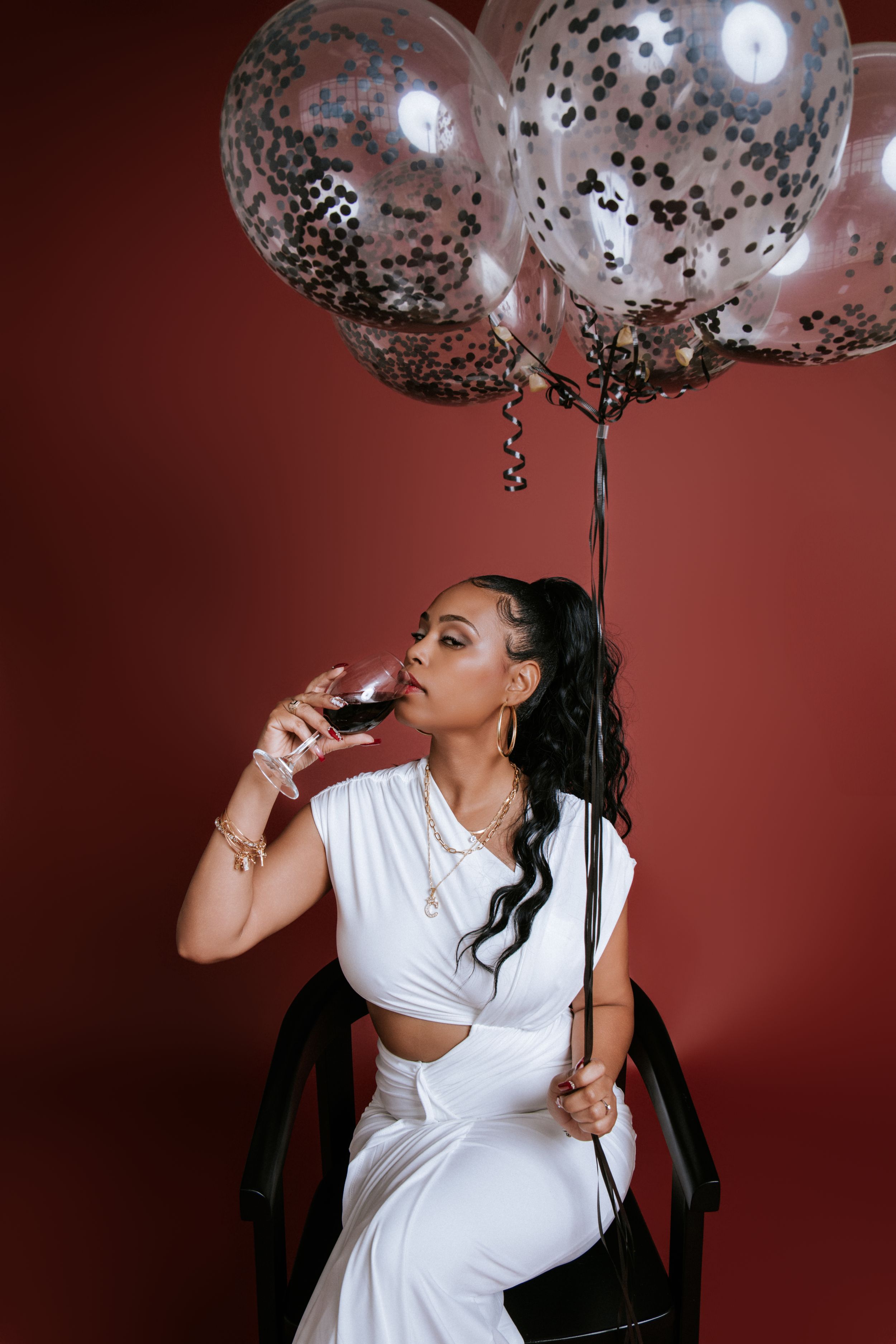 A figure in a white dress poses against a burgundy backdrop with black and silver confetti balloons.