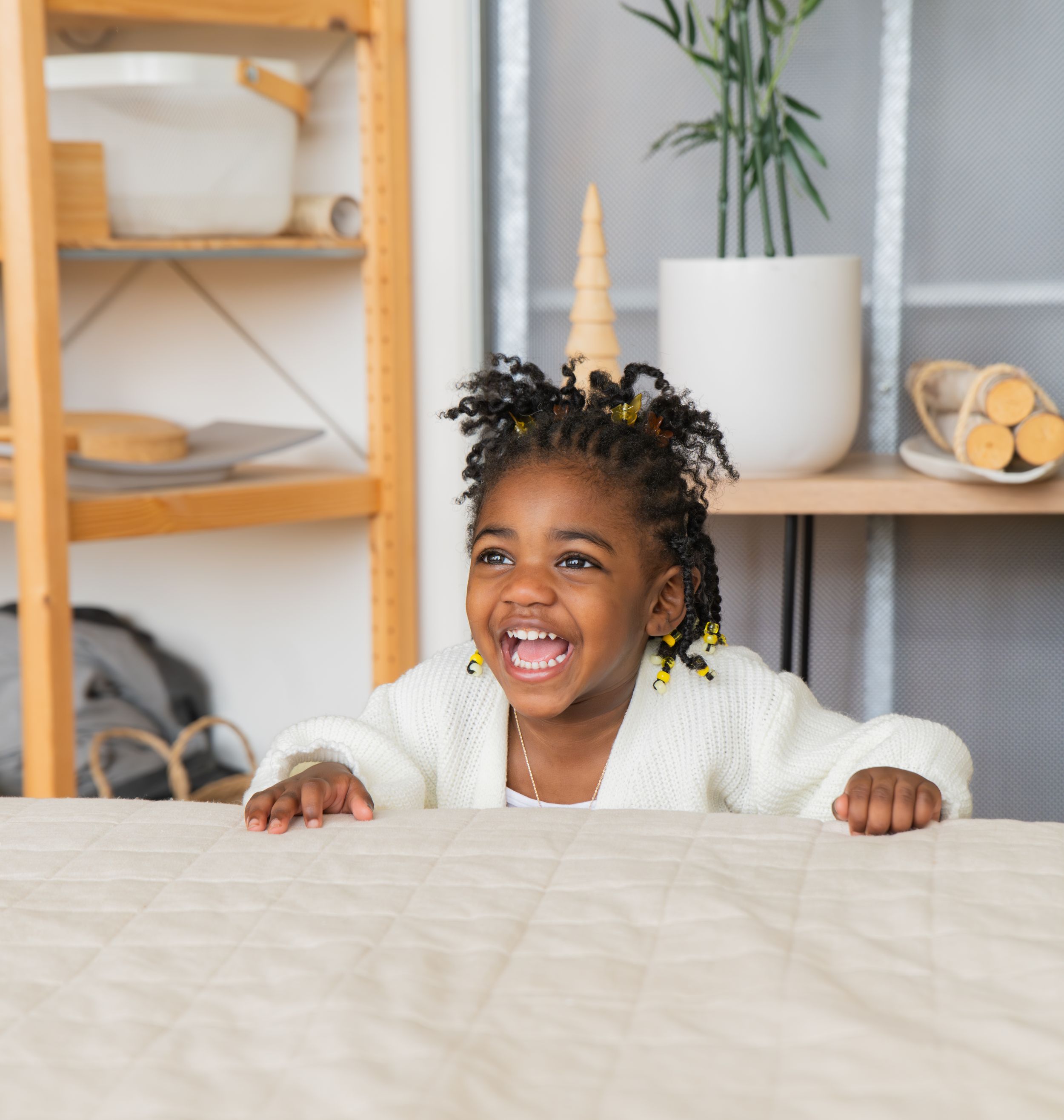 A joyful expression peeks over the edge of a white bedspread in a cozy bedroom with wooden shelving and decor.