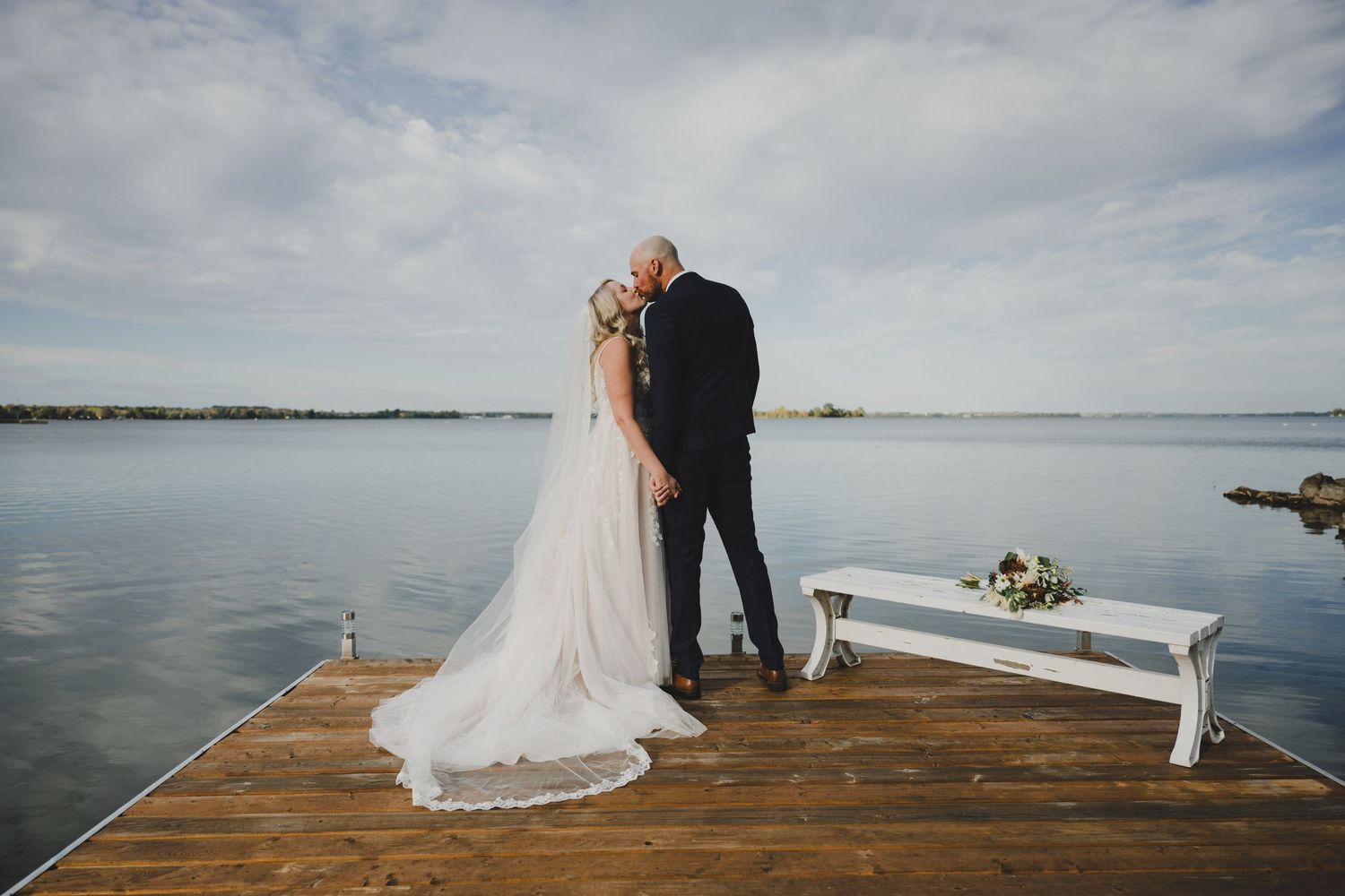 A couple in wedding attire walks through a scenic garden path surrounded by blooming white flowers and greenery.