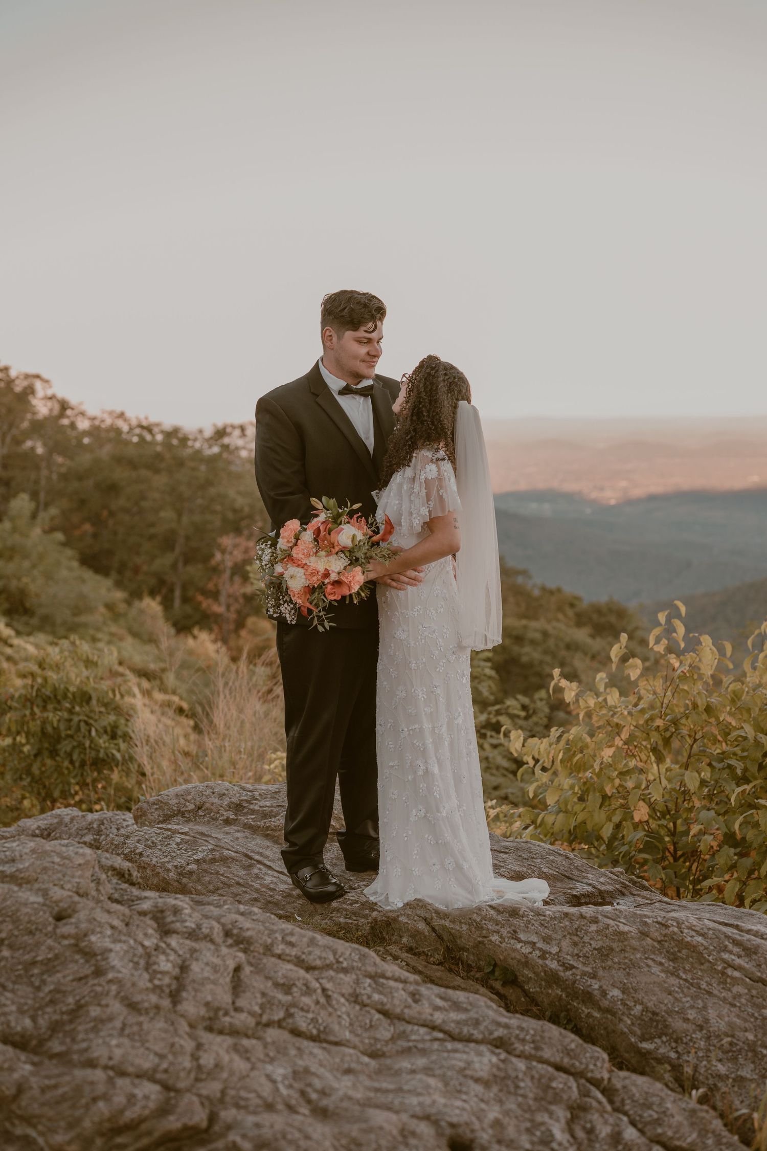 A romantic couple in wedding attire embraces on a rocky mountain overlook during sunset.