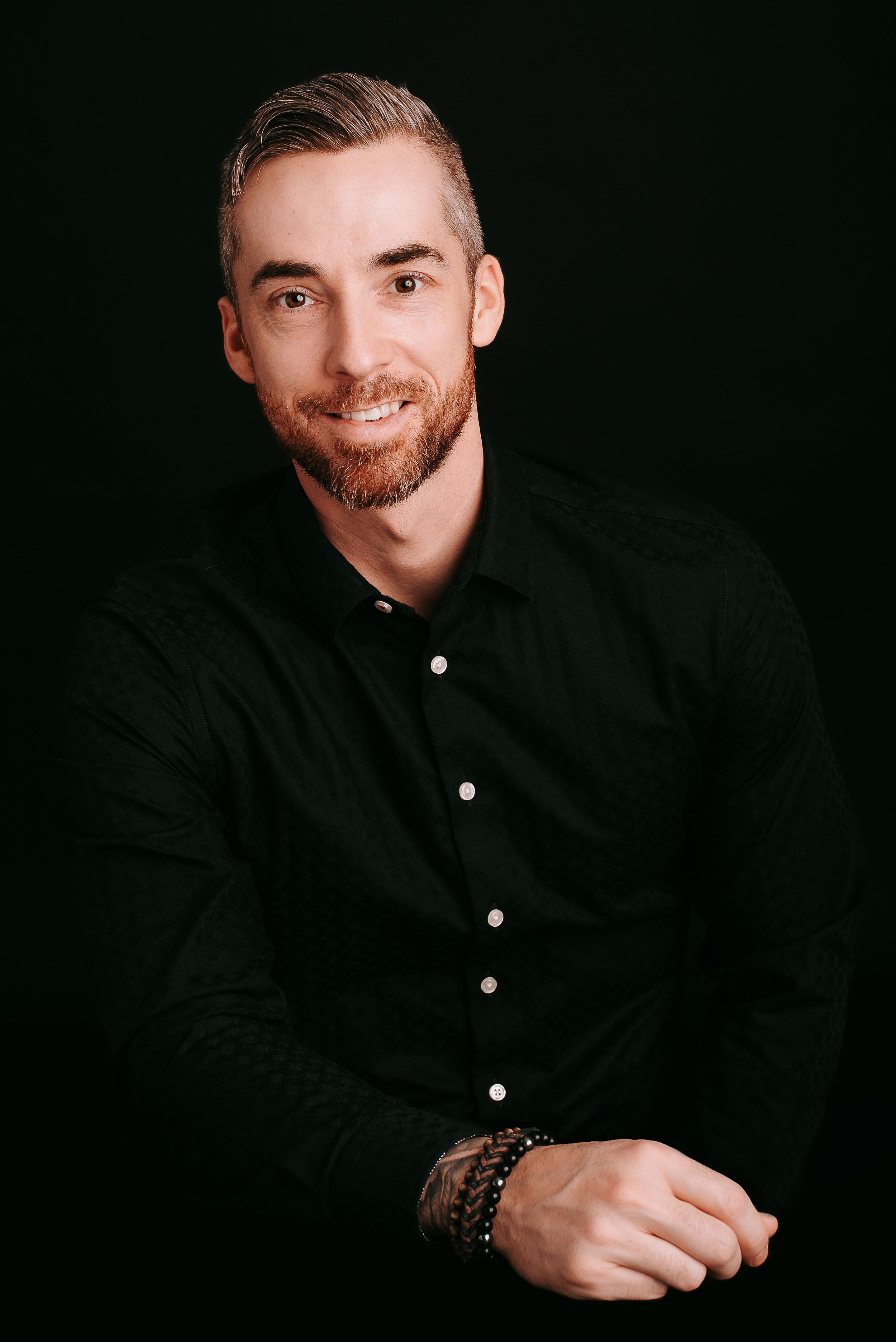 Professional headshot of man in black button-up shirt against dark background.