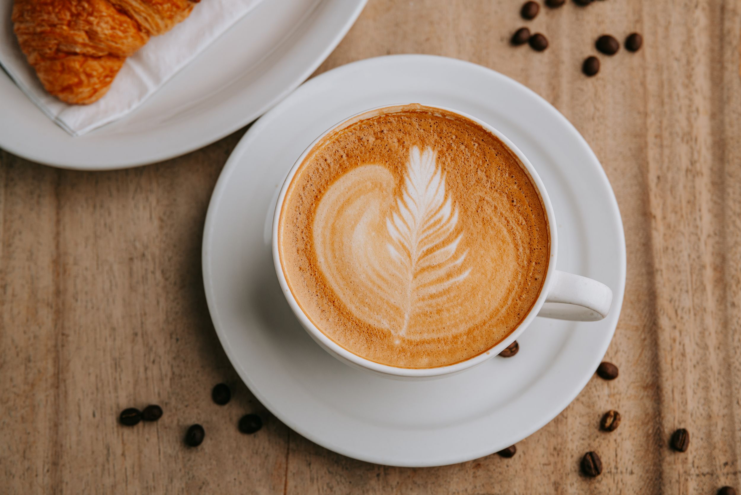 Latte art featuring a leaf design in coffee foam surrounded by scattered coffee beans on a wooden surface.