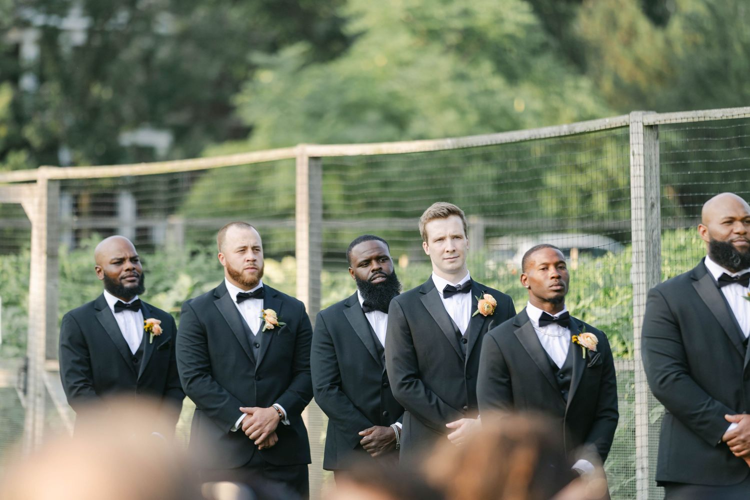 A row of groomsmen in black tuxedos stand together outdoors during a wedding ceremony. - Elawa Farm