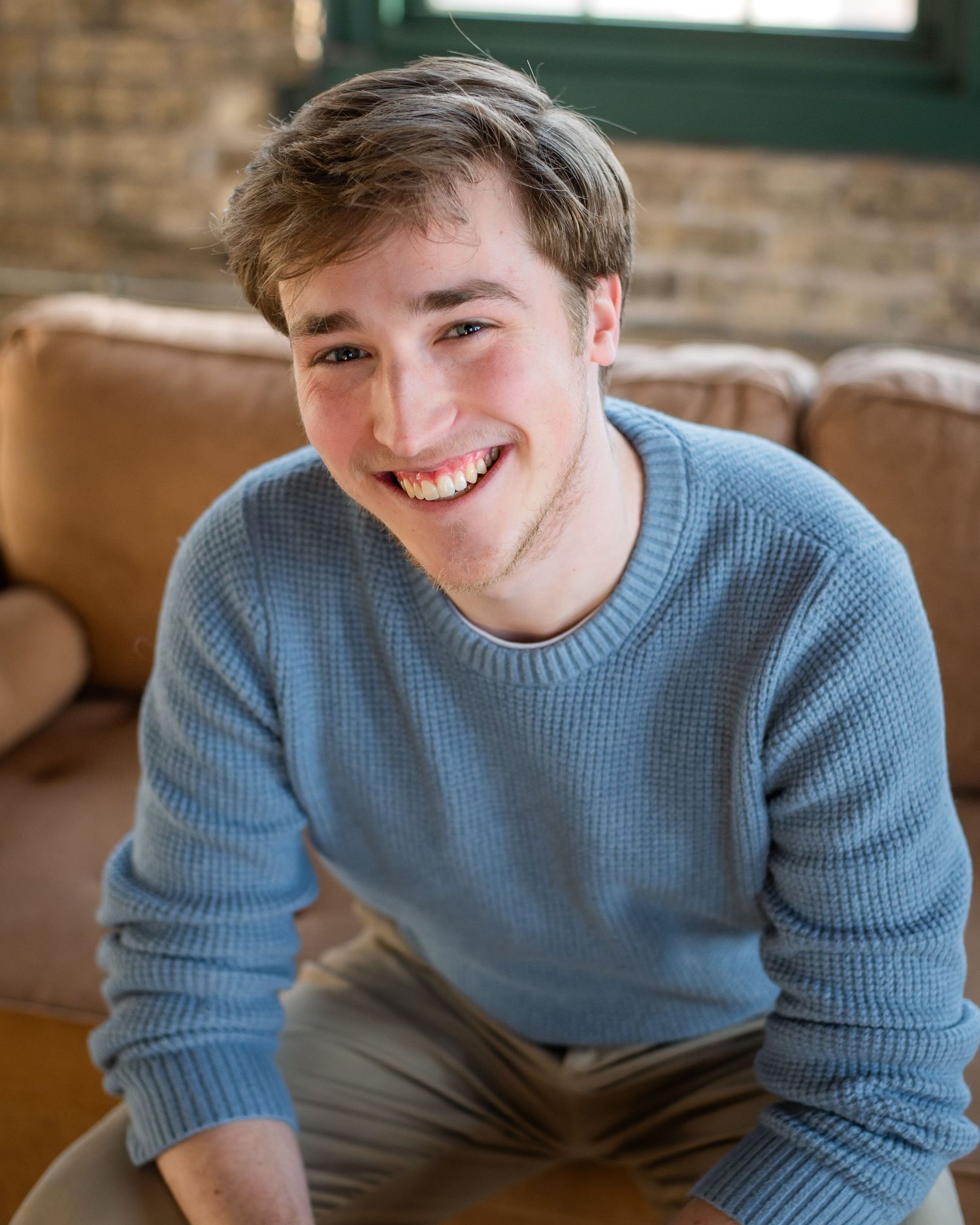 A smiling figure in a light blue sweater sits casually in a warm-lit interior setting.