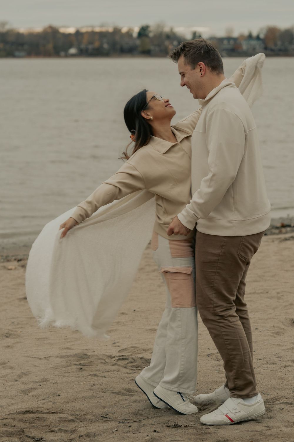 A couple in neutral tones dances playfully on a beach with water and cloudy skies in the background.