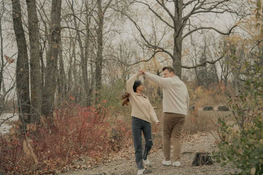 A couple walks hand in hand through a misty autumn forest with bare trees and fallen leaves on the ground.